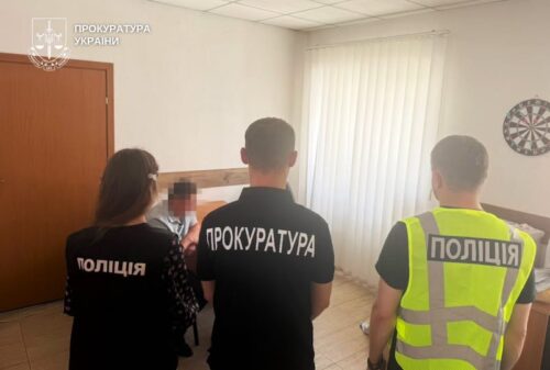 Three officers stand inside a room: a prosecutor in a black shirt reading 'ПРОКУРАТУРА' faces away, flanked by a police officer in a black 'Поліція' shirt and another officer in a bright vest marked 'Поліція'. A blurred person is present near them.