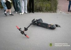 Electric scooter lies on an asphalt road after an incident while bystanders watch; police logo appears in the corner.