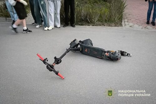 Electric scooter lies on an asphalt road after an incident while bystanders watch; police logo appears in the corner.