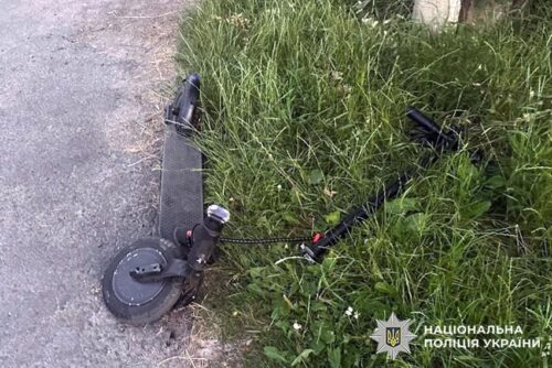 Electric scooter lying on its side on a grass verge beside a road, with a police watermark in the corner.