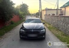 Black BMW sedan with front-end damage parked on a narrow rural street, facing the camera; police watermark bottom right.