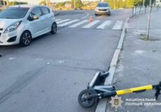 Folded kick scooter lying on the curb near a crosswalk at an urban intersection; a white car and another vehicle in the background with an orange cone nearby.