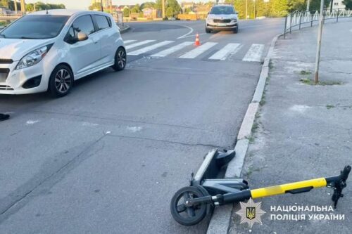 Folded kick scooter lying on the curb near a crosswalk at an urban intersection; a white car and another vehicle in the background with an orange cone nearby.