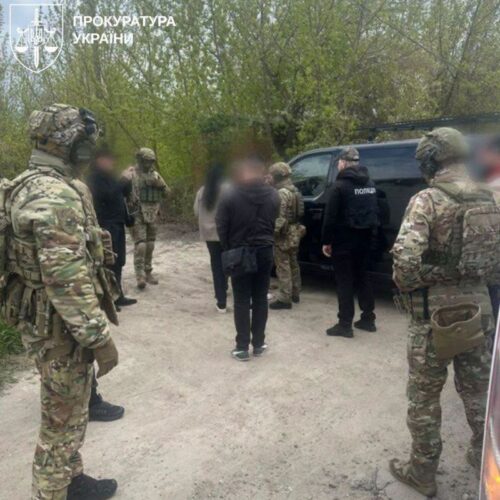 Armed soldiers and police stand around two civilians next to a dark van in a dirt clearing; прокурaтура України watermark in the corner.