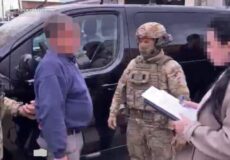 Uniformed soldier with helmet talks to a man in a blue sweater beside a black van; another person with a clipboard records details, all faces pixelated.