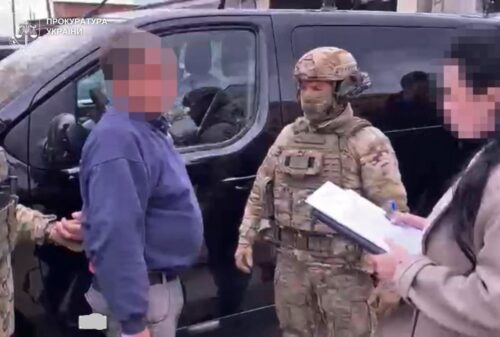 Uniformed soldier with helmet talks to a man in a blue sweater beside a black van; another person with a clipboard records details, all faces pixelated.
