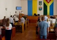 Audience stands in a council chamber as a speaker delivers a briefing from a podium; Ukrainian flag and other banners hang behind the stage, with several attendees wearing Red Cross vests.