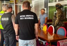 Group of Ukrainian police and prosecutors interview a man at a table inside a building; officers wear black shirts with city text, others in vests or camo nearby.