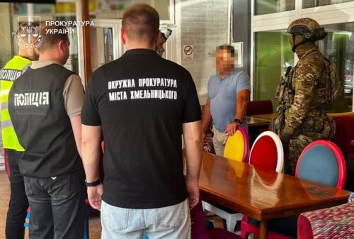 Group of Ukrainian police and prosecutors interview a man at a table inside a building; officers wear black shirts with city text, others in vests or camo nearby.