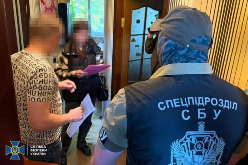 SBU special forces officer in blue jacket questions two blurred suspects in a doorway hallway during a raid.