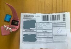 Pink children's watch with the strap open, placed next to a printed shipping label with a barcode on a wooden table.