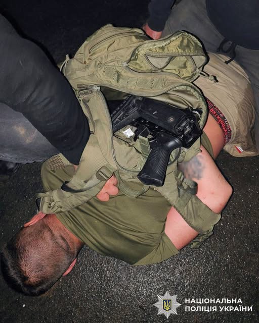 Person lying on a road with an open backpack containing a black handgun; police insignia visible in the corner.