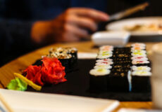 Plate of assorted sushi rolls with pickled ginger and wasabi on a black platter; a person with chopsticks sits in the background.