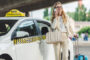 Woman in a light suit stands beside a white taxi, talking on her phone, with a beige bag and rolling blue suitcase in hand.