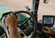 Farmer's hands on tractor steering wheel inside cab, with GPS guidance screen and dusty field ahead.