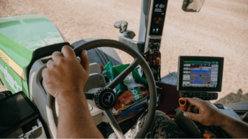 Farmer's hands on tractor steering wheel inside cab, with GPS guidance screen and dusty field ahead.