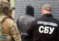 SBU counterintelligence officer in a black jacket labeled Контррозвідка СБУ detaining a person in a gray hoodie against a brick wall background.