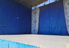 Empty outdoor stage with blue curtains and patterned vertical banners, a raised wooden platform under an open roof.