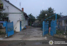 Open blue metal gates lead into a paved courtyard between houses on a cloudy day, with a small garden and clothesline visible; National Police of Ukraine watermark in the bottom right.