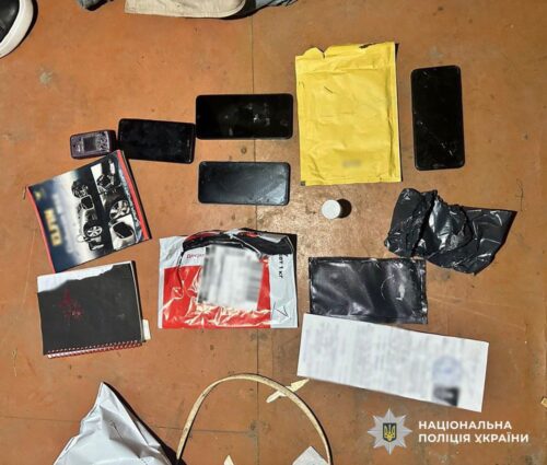 Several smartphones, a yellow padded envelope, papers, and small bags laid out on a brown floor, with a Ukrainian police logo in the corner.