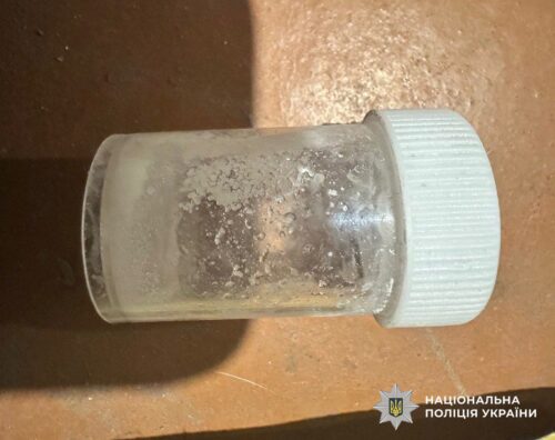 Transparent plastic vial with a white screw cap on a brown surface; dusty residue inside.
