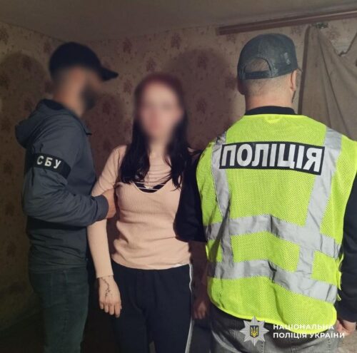 Two police officers detain a woman indoors; one wears a neon vest marked 'ПОЛІЦІЯ' and stands beside them.