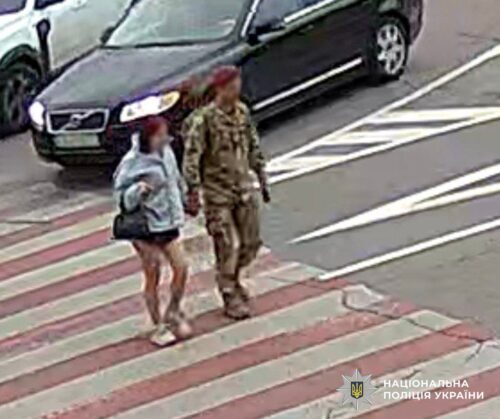A soldier in camouflage walks beside a woman with a handbag across a striped crosswalk near parked cars.