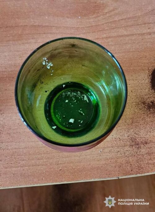Top-down view of a green glass with a few white residues at the bottom on a wooden table; police watermark in the corner.