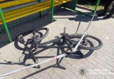 Black bicycle lying on its side on a sunlit sidewalk, cordoned off with police tape near a bench.