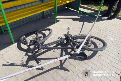 Black bicycle lying on its side on a sunlit sidewalk, cordoned off with police tape near a bench.