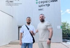 Two men stand in front of a metal-clad service center building with the sign 'Сервісний центр' in Cyrillic, on a sunny day.