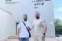 Two men stand in front of a metal-clad service center building with the sign 'Сервісний центр' in Cyrillic, on a sunny day.