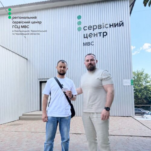 Two men stand in front of a metal-clad service center building with the sign 'Сервісний центр' in Cyrillic, on a sunny day.