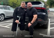 Two men in black athletic shirts pose beside a dark SUV in a parking lot; one has a prosthetic leg and uses a crutch, both looking at the camera.