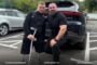 Two men in black athletic shirts pose beside a dark SUV in a parking lot; one has a prosthetic leg and uses a crutch, both looking at the camera.