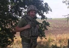 A soldier in camouflage uniform and helmet standing in a field beside a tree, adjusting his tactical vest.