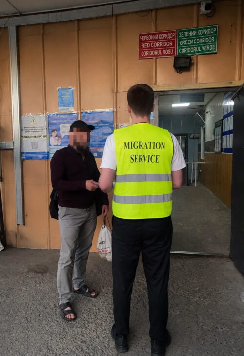 Migration Service officer in a neon vest speaks with a man outside an office entrance, handing him a bag.