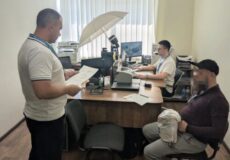 Office scene: two employees at a desk with paperwork, one standing and reading documents while another works at a computer; a large umbrella light sits above the desk.