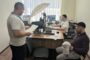 Office scene: two employees at a desk with paperwork, one standing and reading documents while another works at a computer; a large umbrella light sits above the desk.