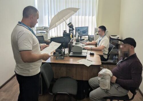 Office scene: two employees at a desk with paperwork, one standing and reading documents while another works at a computer; a large umbrella light sits above the desk.
