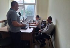 Three men in a small office; one standing at a desk with papers and a microphone, while two sit at computers nearby.