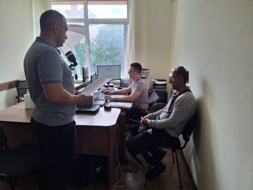 Three men in a small office; one standing at a desk with papers and a microphone, while two sit at computers nearby.