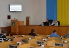 Government meeting in a hall with a blue-yellow Ukrainian flag backdrop and crest; two panelists at the front desk facing seated attendees.
