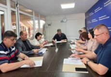 Group of people around a long conference table reviewing papers in a meeting room with a clock on the wall.