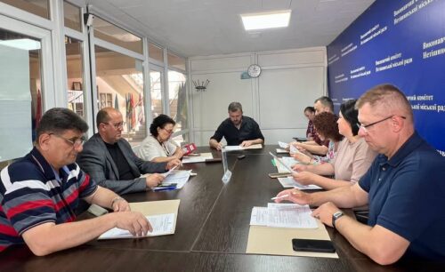 Group of people around a long conference table reviewing papers in a meeting room with a clock on the wall.