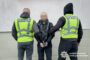 Two Ukrainian police officers in neon vests detain a man with a blurred face against a gray wall; police insignia visible.
