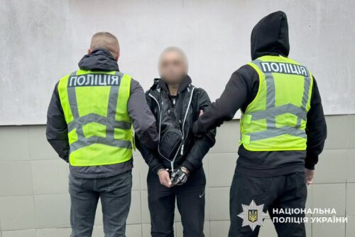 Two Ukrainian police officers in neon vests detain a man with a blurred face against a gray wall; police insignia visible.