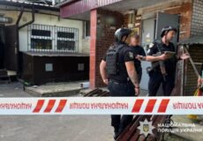 Three Ukrainian police officers in tactical vests outside a building, behind red-and-white caution tape; one holds a tablet.