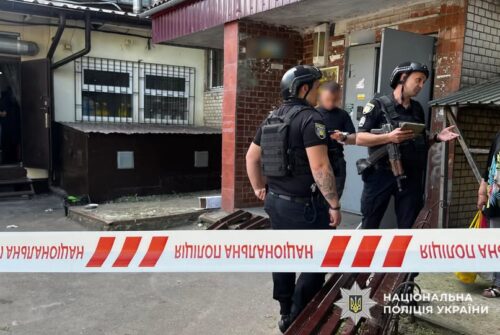 Three Ukrainian police officers in tactical vests outside a building, behind red-and-white caution tape; one holds a tablet.