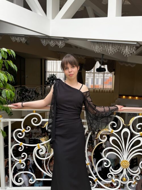 Woman in a black, one-shoulder evening gown leaning on a white ornate balcony railing inside a venue with chandeliers.